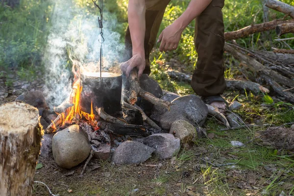 Man's hands are putting firewood into the bonfire, over which hangs a pot with a preparing dinner, on an open fire, on open air on a summer day. Tourism and recreation.
