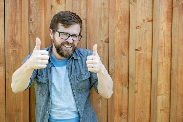 一个快乐的 留着胡子的戴眼镜的男人用双手竖起大拇指 微笑着看着摄像机 背对着一堵旧木墙 后续行动 — 图库照片