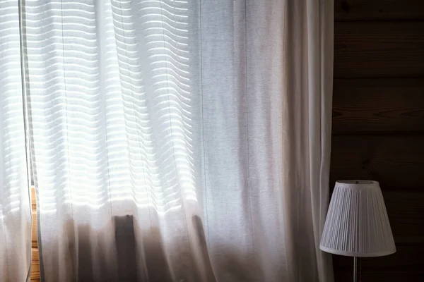 窗户上透出透明的窗帘 透过窗帘可以看到阳光 可以看到花环的影子 旁边是一盏灯 靠着一堵木制的墙 在一个舒适的卧室里 — 图库照片