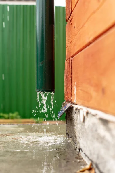 Rain water flows from a drainpipe to a concrete foundation, a wooden house, against a green fence. Close-up. — Stock Image Rain Water Flows Drainpipe Concrete Foundation Wooden House Green Fence Royalty Free Stock Photos