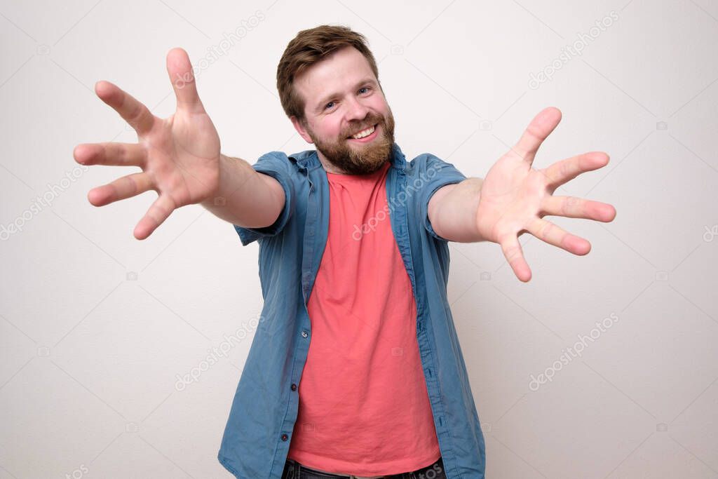 A good-natured bearded man pulls his hands to the camera and wants to cuddle, smiling widely welcoming a friend. Isolated on white background. Close-up.