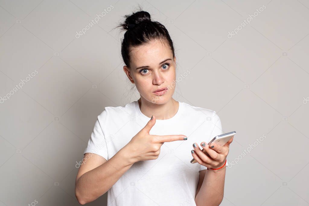 Young, pretty woman holds a smartphone in her hands and points with her index finger at it. She is confused by what she saw or read on the phone and looks inquiringly at the camera. Isolated on white background