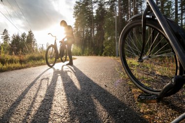 Yolun kenarında duran eski bir bisikletin tekerleği, arka planda, bir yaz akşamında güneşin batışında bisiklete binecek olan bir kadının silüeti..
