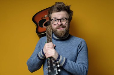 Eski süveter giymiş komik bir hippi akustik bir gitarı omuzunda tutuyor ve tuhaf bir suratla kameraya bakıyor..