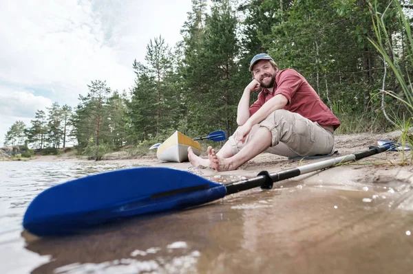 Glad leende man vilar på stranden av sjön efter att ha rest med kajak. Livsstil. — Stockfoto