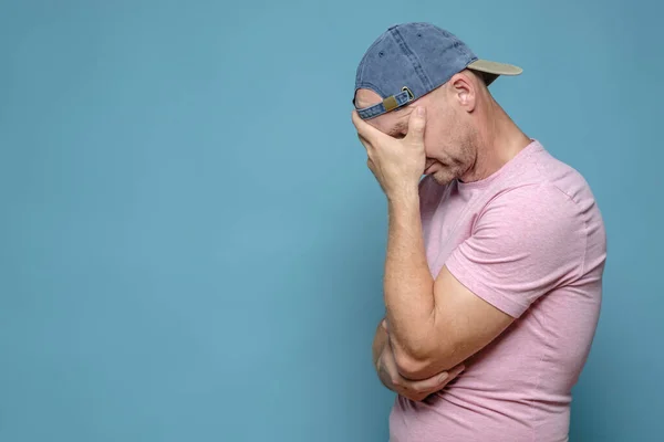 Der Mann mit Mütze und T-Shirt war sehr müde, aufgebracht und gestresst, das Gesicht mit der Hand bedeckt und den Kopf gesenkt. Kopierraum. Blauer Hintergrund. — Stockfoto