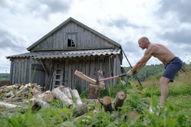 Güçlü bir adam çıplak gövdesiyle köyde büyük bir çelik baltayla odun kesiyor, eski bir ahırın arka planında..