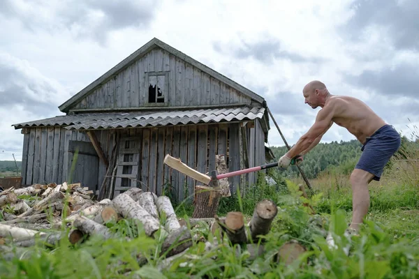 Güçlü bir adam çıplak gövdesiyle köyde büyük bir çelik baltayla odun kesiyor, eski bir ahırın arka planında..