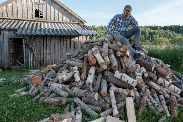 Pensive člověk sedí na hromadě dříví, je unavený a neví, jak štípnout klády. Venkovský životní styl. — Stock fotografie