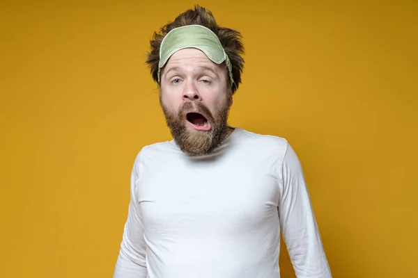 Shaggy, bearded man with a sleeping mask on his head yawns amusingly. Yellow background. — Stock Photo, Image