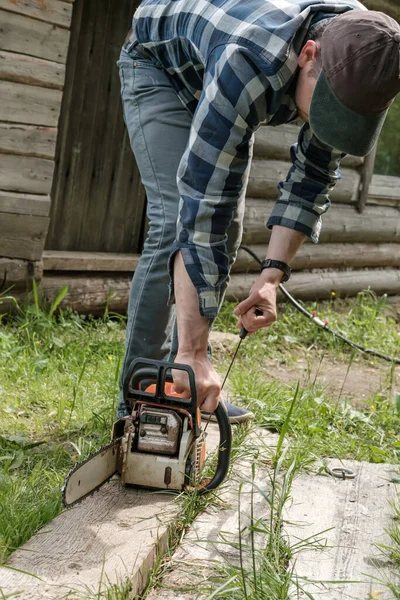 Homem focado prepara uma serra profissional a gasolina para o trabalho, contra o pano de fundo de um antigo celeiro de madeira no campo. — Fotografia de Stock