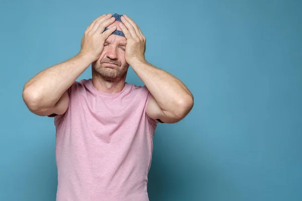 Измученный, страдающий белый мужчина держит голову руками, от стресса или головной боли. Принято. Синий фон. — стоковое фото