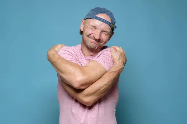 If you are lonely, hug yourself. Cute man in a cap and a pink T-shirt hugs himself and dreams with eyes closed. Blue background. — Stock Photo, Image