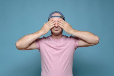 Şapkalı ve pembe tişörtlü sakin bir adam elleriyle gözlerini kapadı. Bir sürpriz bekliyor ya da bir şey görmek istemiyor.. 