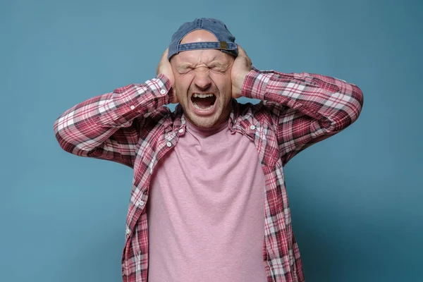 De mens staat onder stress, is moe van het lawaai, bedekt oren met handen en schreeuwt zenuwachtig. Blauwe achtergrond. — Stockfoto