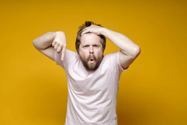 Disgruntled, strange man points down with index finger, holds his hair with hands and looks seriously while saying something. — Stock Photo, Image