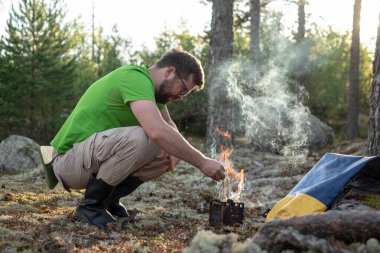 Yürüyüş kıyafetli bir adam, ormanda ve kampta yemek pişirmek için prefabrik ocağı odunla ısıtıyor. Etkin dinlenme.