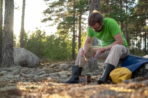 Caucasian man in hiking clothes uses wood to heat a metal burner for cooking, in the forest, in a camp, on a summer evening.