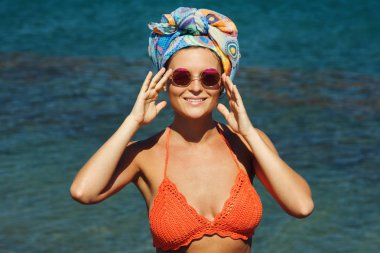 Kumsalda güneş gözlüğü ve başörtüsü giyen kadın turuncu örgü bikini,