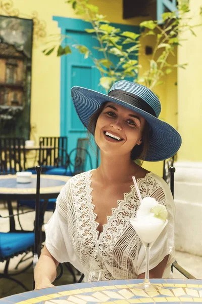 Happy woman wearing blue hat is enjoying her cold cocktail in city cafe