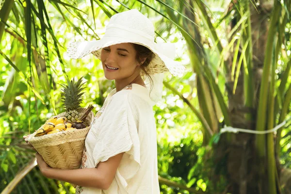 新鮮なフルーツのバスケットとトロピカル ガーデンの広いつばの帽子を着ている美しい女性 — ストック写真