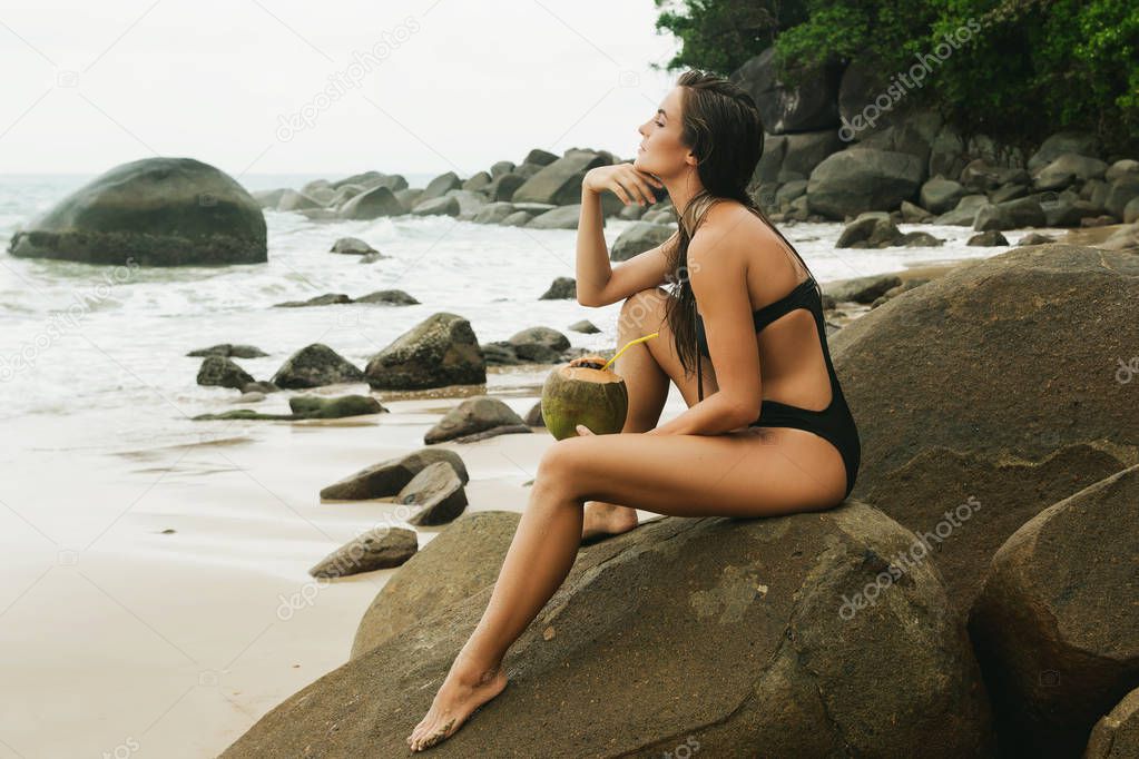 Woman sitting on the rocky beach is enjoy her coconut drink, and listen sound of the sea