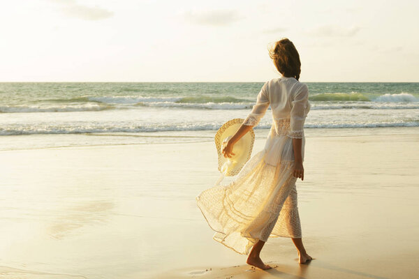 Счастливая молодая женщина в красивом белом платье, гуляющая по пляжу на закате
