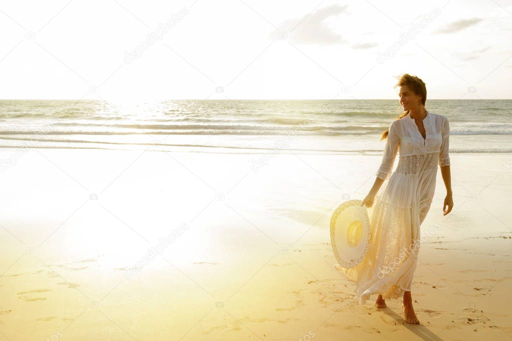 Happy young woman wearing beautiful white dress walking on beach during sunset