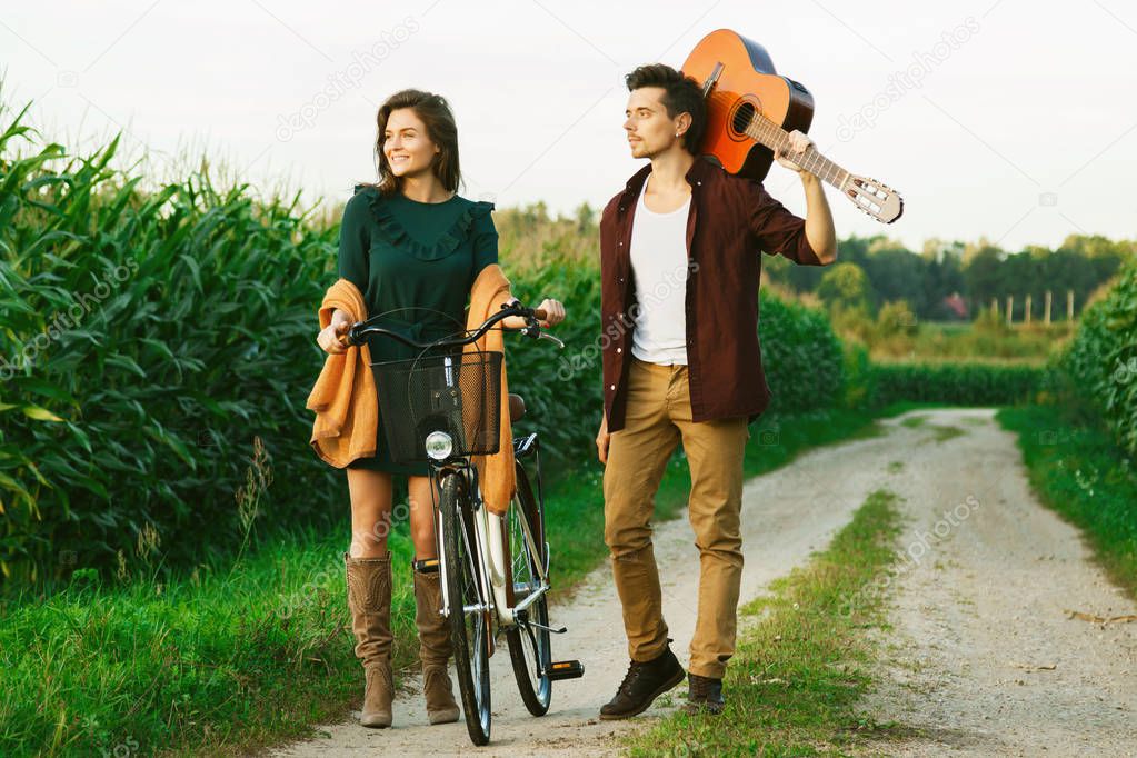 Young couple walking on country road. Girl riding bicycle and guy is holding guitar.