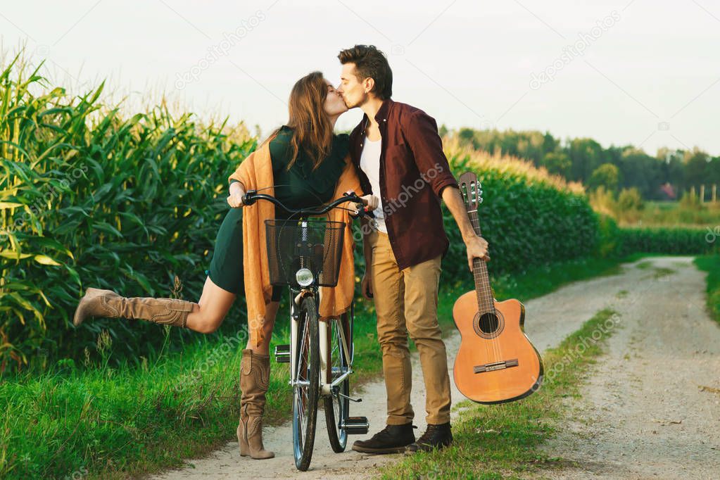 Young couple walking on country road. Girl riding bicycle and guy is holding guitar.