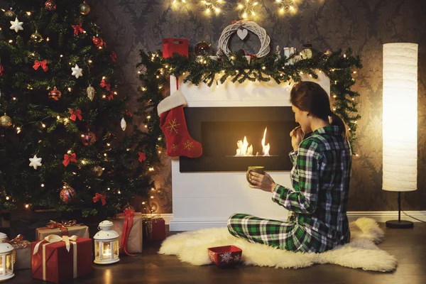 クリスマスや新年のお祝い 暖炉のそばに座っている女性は クッキーと紅茶を飲んでいます — ストック写真