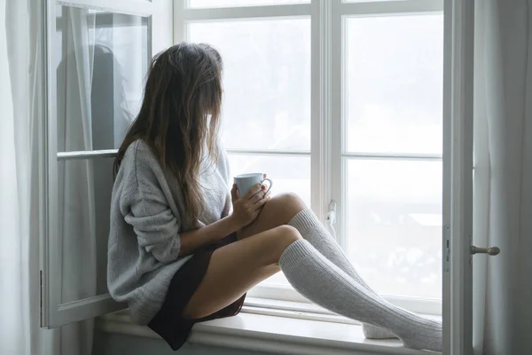 Woman is sitting on the windowsill and drinking hot tea or coffee at snowy winter day