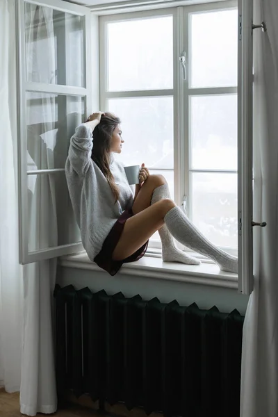 Woman is sitting on the windowsill and drinking hot tea or coffee at snowy winter day