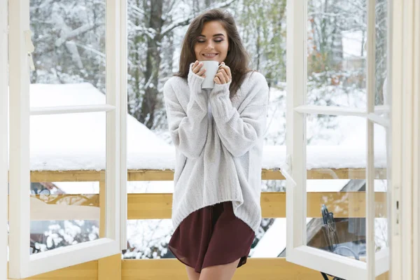 Happy woman with a cup of hot drink is standing on the balcony at snowy winter day