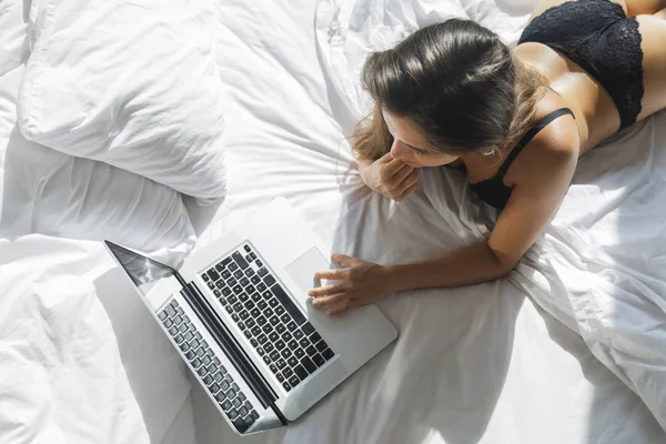 Сексуальна жінка в чорній білизні лежить на ліжку і використовує ноутбук — стокове фото