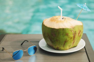 Yüzme havuzunun yanındaki masada taze hindistan cevizi içeceği ve güneş gözlüğü var.