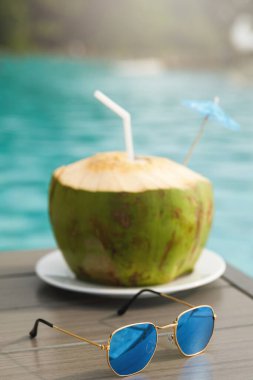 Yüzme havuzunun yanındaki masada taze hindistan cevizi içeceği ve güneş gözlüğü var.