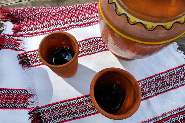 Kvass em copos de barro e jarro em uma mesa de madeira com têxteis nacionais. Autenticidade ucraniana estilo nacional — Fotografia de Stock