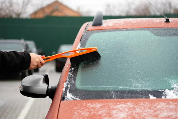 Man clears snow from icy windows of car. Brush in mans hand. Windshield of orange auto, horizontal view.