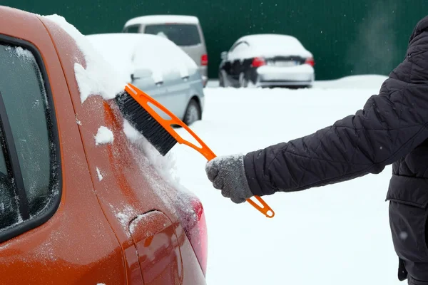 Brush in mans hand. Man clears orange car from snow. Car covered with snow. Winter problems of car drivers. — Stock Photo, Image