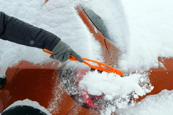 Man clears orange car from snow on a cold winter day after snowfall. Brush in mans hand. Lot of snow on car. — Stock Photo, Image