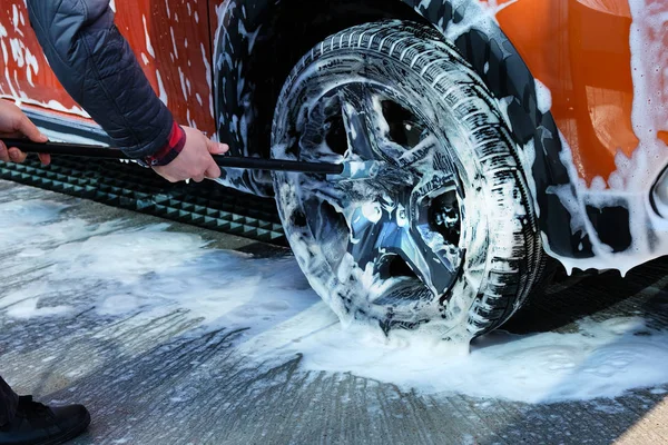 Man washes black wheel of his orange car with brush. Cleaning with soap suds at self-service car wash. Soapy water runs down. — Stock Photo, Image