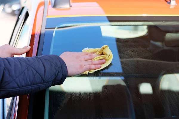 L'uomo dopo lavaggio pulisce il parabrezza della sua auto arancione con uno straccio giallo all'atto di autolavaggio. Primo piano corpo mano e auto maschile . — Foto Stock