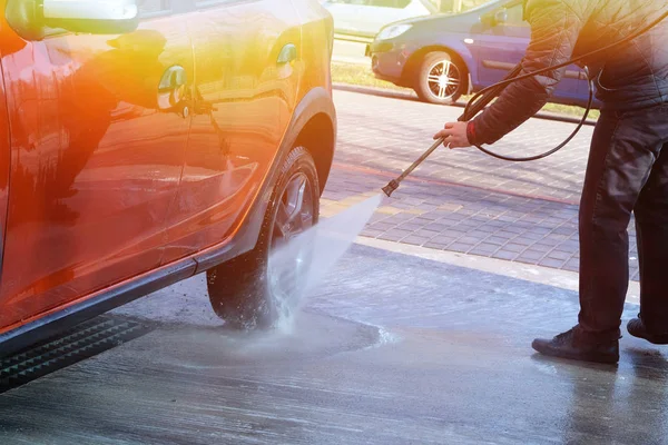 O homem lava o carro laranja na lavagem de carros ao ar livre. Limpeza com água na lavagem de carro self-service. Luz solar . — Fotografia de Stock