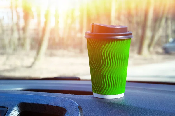 Green paper cup with tea or coffee in car on sunny blurred background. Takeaway, social media. Morning breakfast in auto.