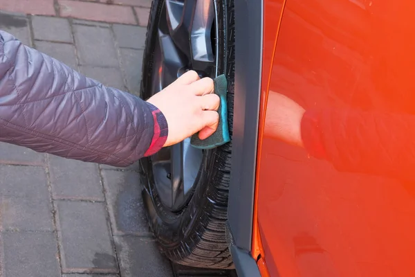 Man after washing applies tire cleaner with a sponge. Orange car at car wash. Male hand and car wheel closeup. — Stock Photo, Image