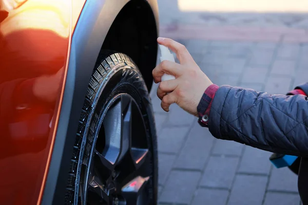 Homem após a lavagem aplica limpador de pneus com um spray branco. Carro laranja na lavagem de carros. Mão masculina e roda de carro de perto . — Fotografia de Stock