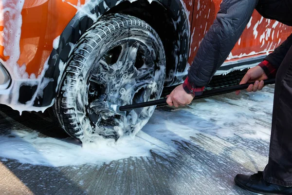 Cleaning with soap suds at self-service car wash. Man washes black wheel of his orange car with brush. Soapy water runs down. — Stock Photo, Image