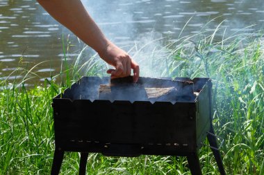 Yakıt ve kömür ile ızgara. El ateşe odun ekliyor. Nehir kenarında barbekü. Güneş.