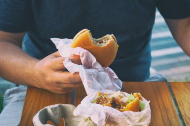 El kafede ahşap masada taze lezzetli burger tutuyor. Sığır eti, domates, peynir, salatalık ve marul ile ızgara burger. Öğle yemeği için fast food. Yumuşak netleme, tonda görüntü.